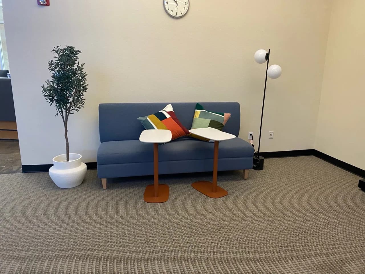 A completed commercial seating area with a blue sofa, two compact tables, a floor lamp, and a potted tree arranged against a clean wall.